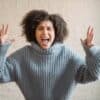 Irritated African American female with raised arms looking at camera while shouting loudly near wall in light room at home
